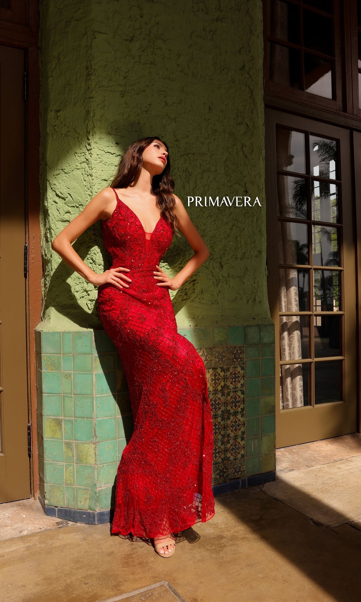 A woman in a fitted, sleeveless red Open-Back Primavera Long Beaded Prom Dress 4113 stands against a green textured wall as sunlight casts shadows;